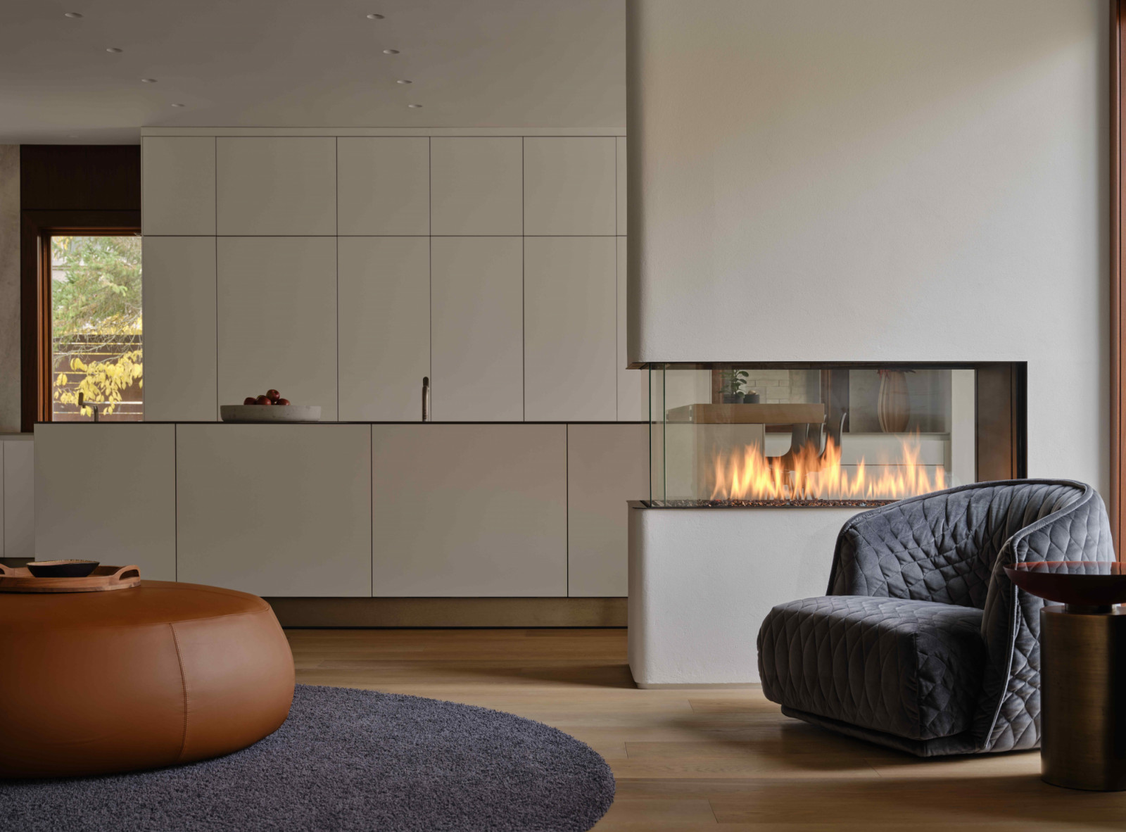 A three-sided glass fireplace housed within a plaster-wrapped pillar subtly divides the seating area from the kitchen