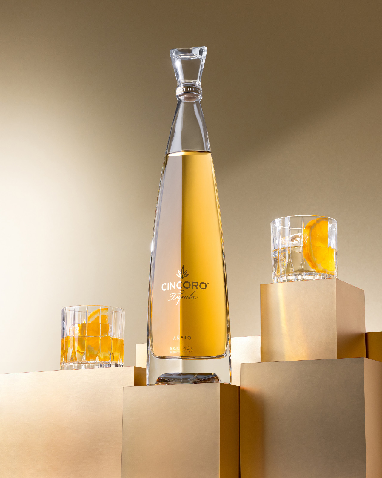 A bottle of premium Cincoro Añejo tequila displayed next to a crystal glass containing a single large ice cube and a fresh orange slice garnish.