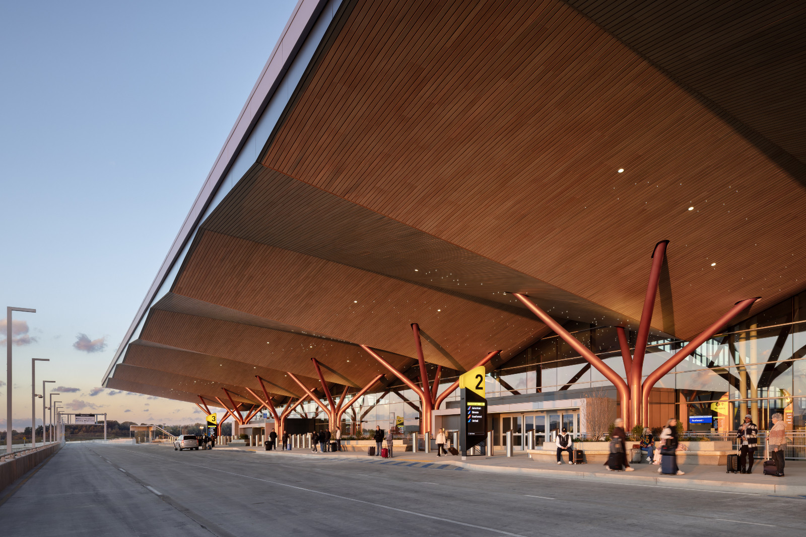 At dusk, branching red steel columns support a deep wood-clad canopy along the departures kerb, their forked forms reading like a row of trees against the evening sky (Photo: Ema Peter)