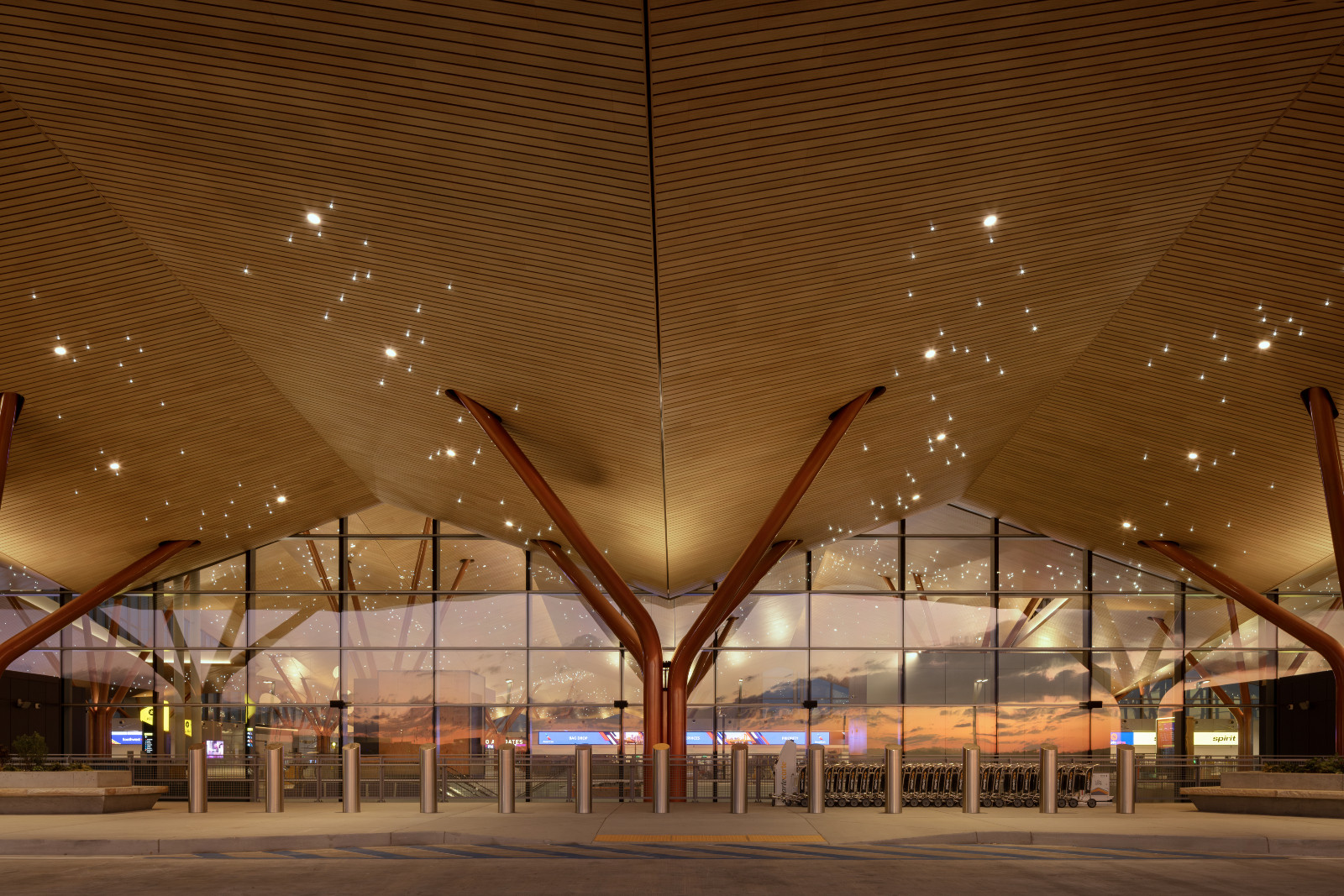 At twilight, the canopy’s branching columns glow against a glass facade, with the wood ceiling’s scattered lights and an orange sunset reflected beyond (Photo: Ema Peter)