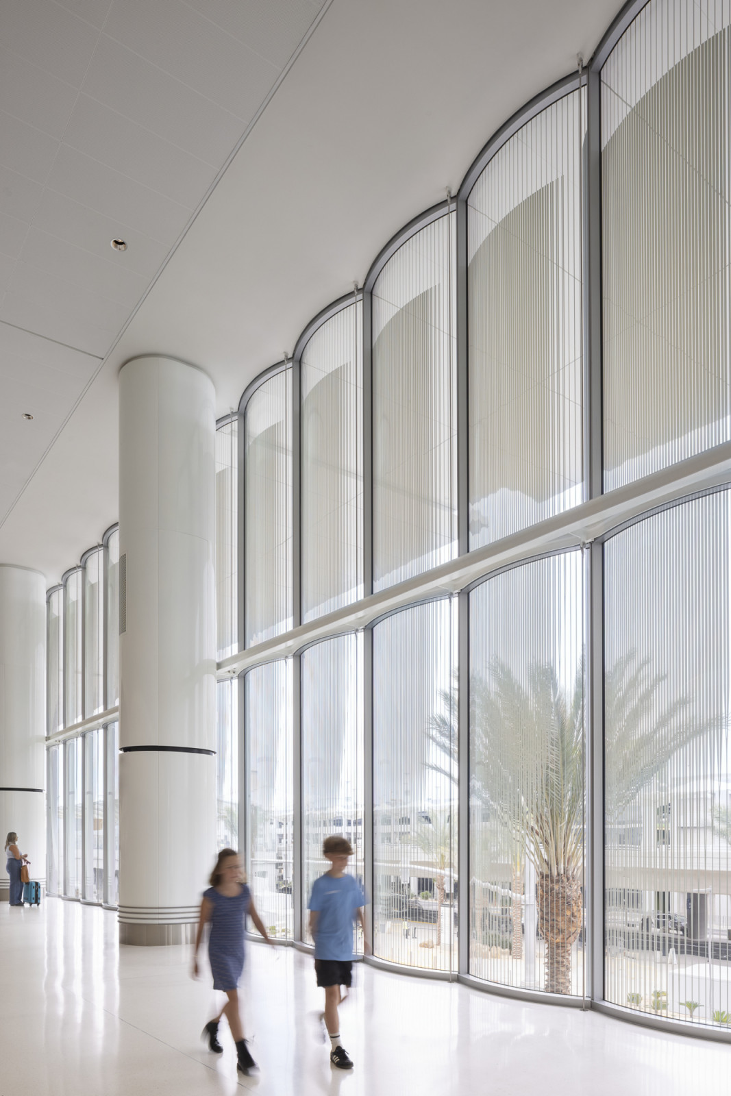 Inside, a procession of tall curved glass bays floods the concourse with daylight, their fluted screens filtering views of palm trees and the airfield beyond (Photo: Ema Peter)