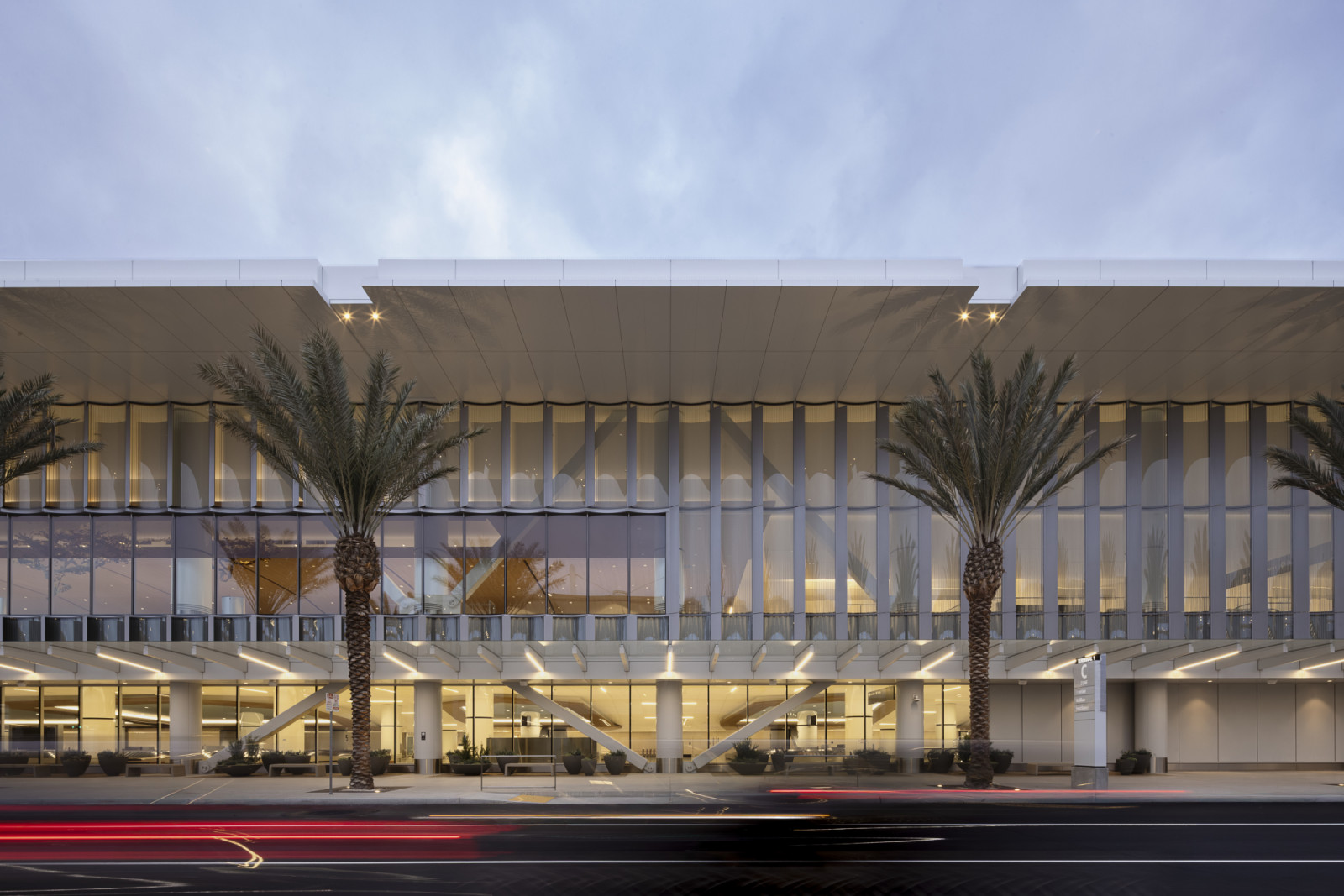 Terminal 1’s curved glass facade catches the evening light, its rippling fluted screen and angled cross-bracing visible behind a line of palm trees (Photo: Ema Peter)