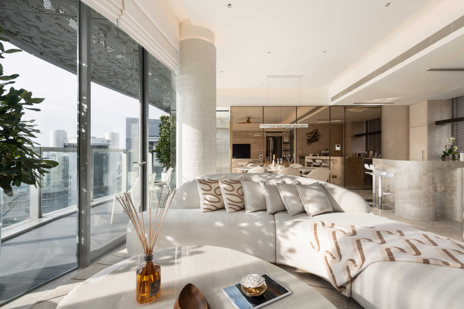 A curved sofa anchors the living area, which is separated from the dining area by a mosaic-clad column, while full-height glazing opens to the balcony and city views beyond
