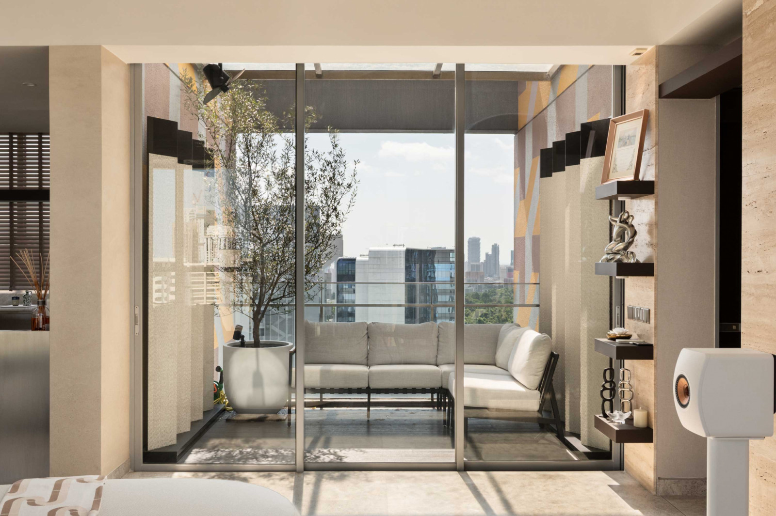 Sliding glass doors extend the living area to the balcony, where an L-shaped settee and potted olive tree frame views of the surrounding cityscape