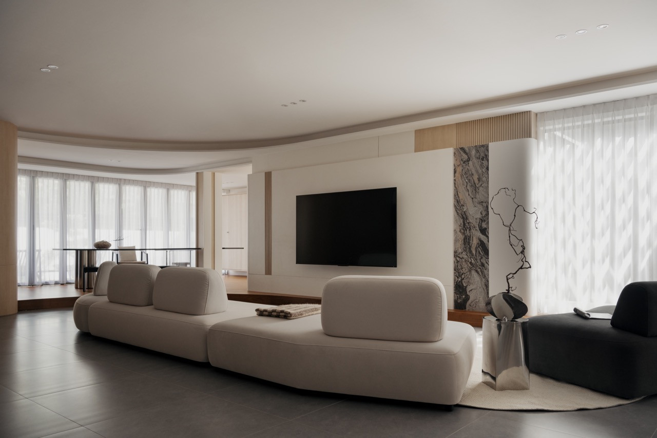 The ground-floor living area with modular cream sofa, curved ceiling, Arabescato Orobico marble feature panel, and the dining platform visible beyond through sheers