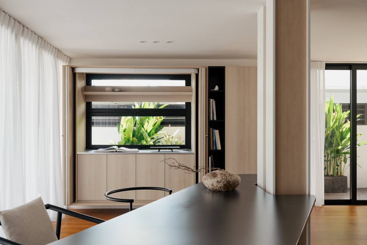 View across the dining table towards the slide-up kitchen window framing garden greenery beyond, with teak flooring and sheer curtains to either side