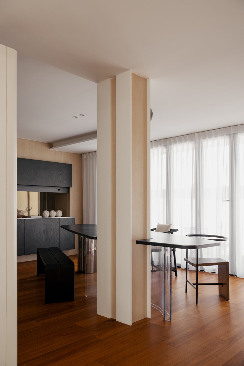 The dining area with the custom organic-form dining table and acrylic-leg seating, the structural column integrated into the composition, with the dark kitchen cabinetry visible beyond