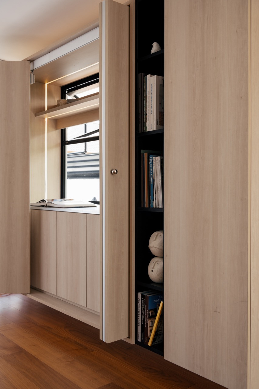 A children’s room with a built-in window bench, oak-toned cabinetry, a slide-up window, and an open bookshelf unit with ceramic objects and design books
