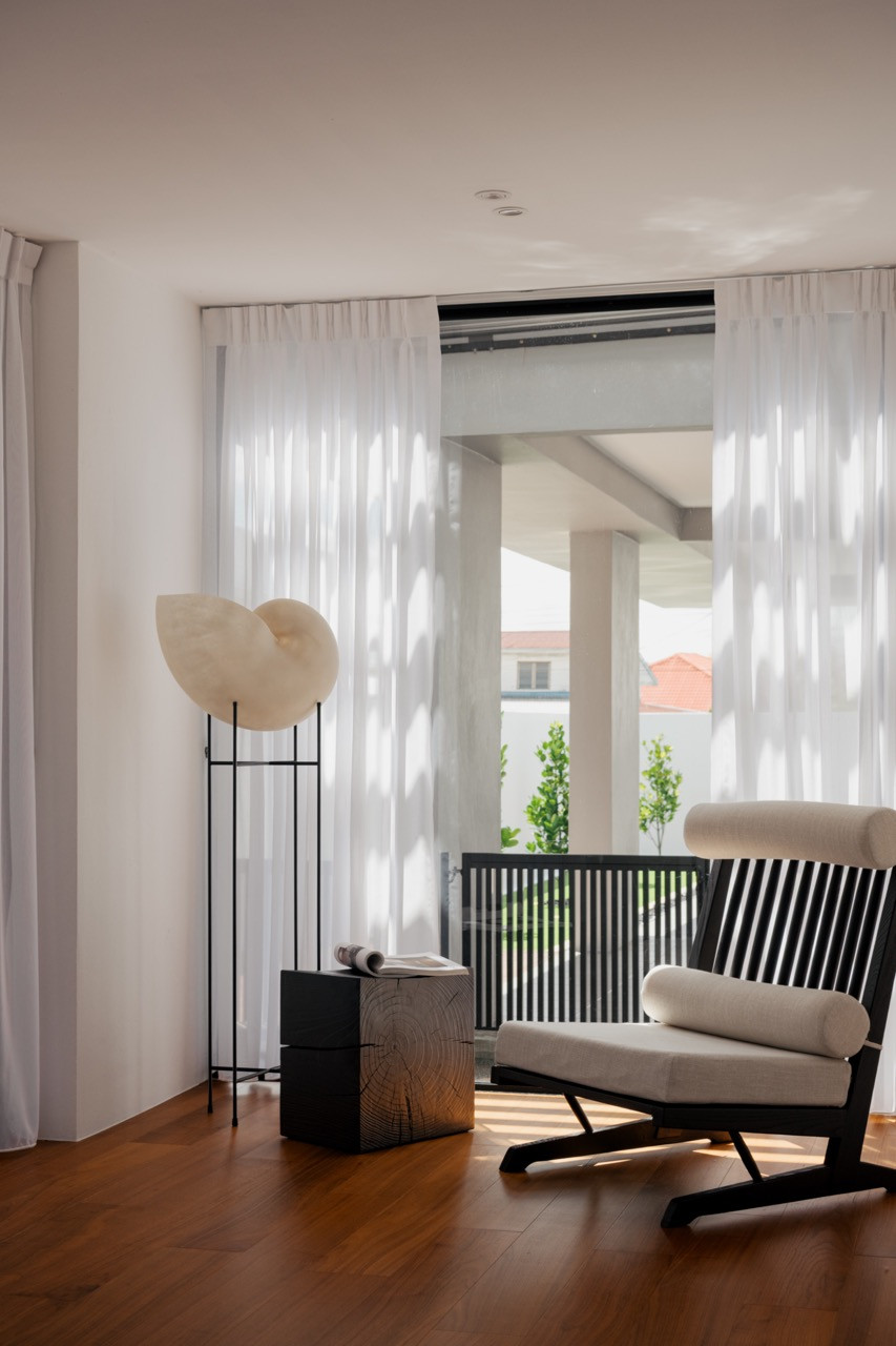 The master bedroom sitting area with teak flooring, a slatted lounge chair, and a sculptural floor lamp beside sheer curtains looking onto a balcony