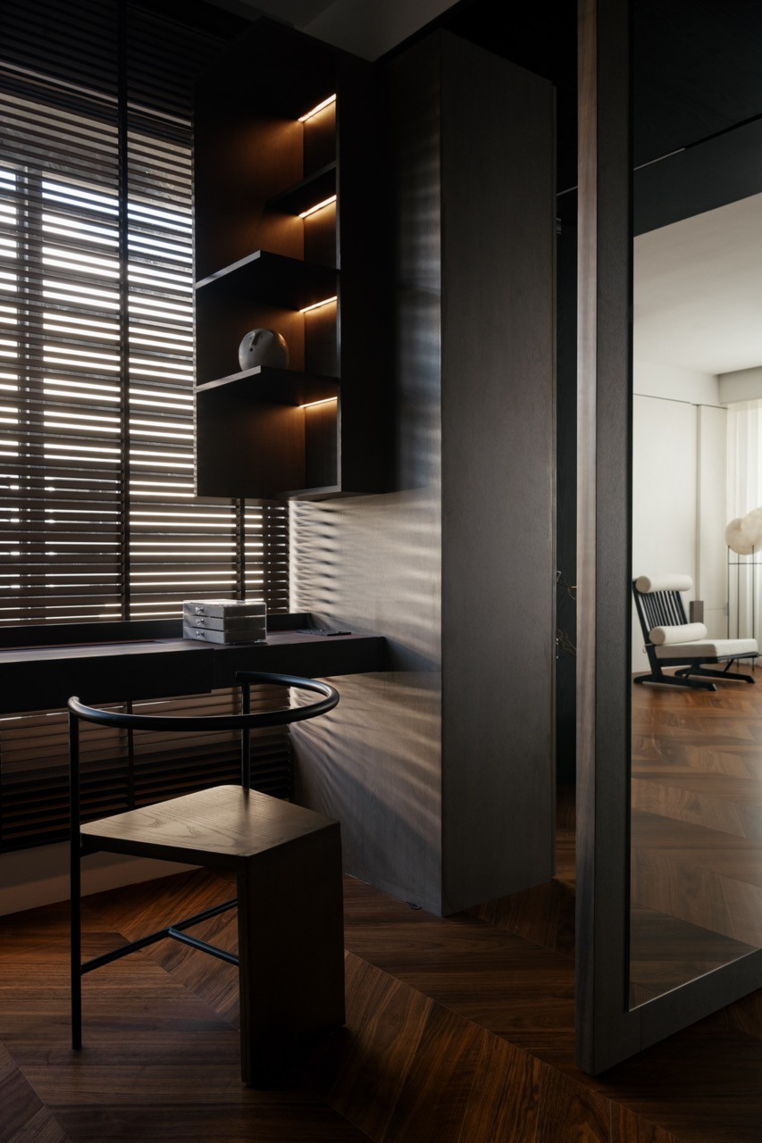 A dark-toned dressing area with built-in shelving and strip lighting opens to the master bedroom beyond, where a slatted lounge chair is visible