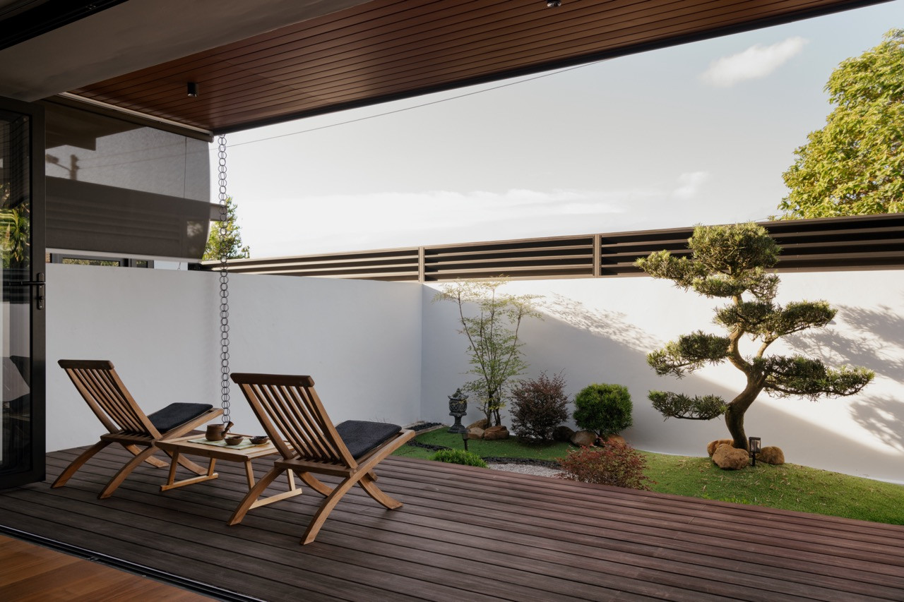 The backyard garden with timber deck, teak loungers, cloud-pruned tree, and landscaped planting bed with river stones and ornamental shrubs, viewed in daylight with the folding doors open