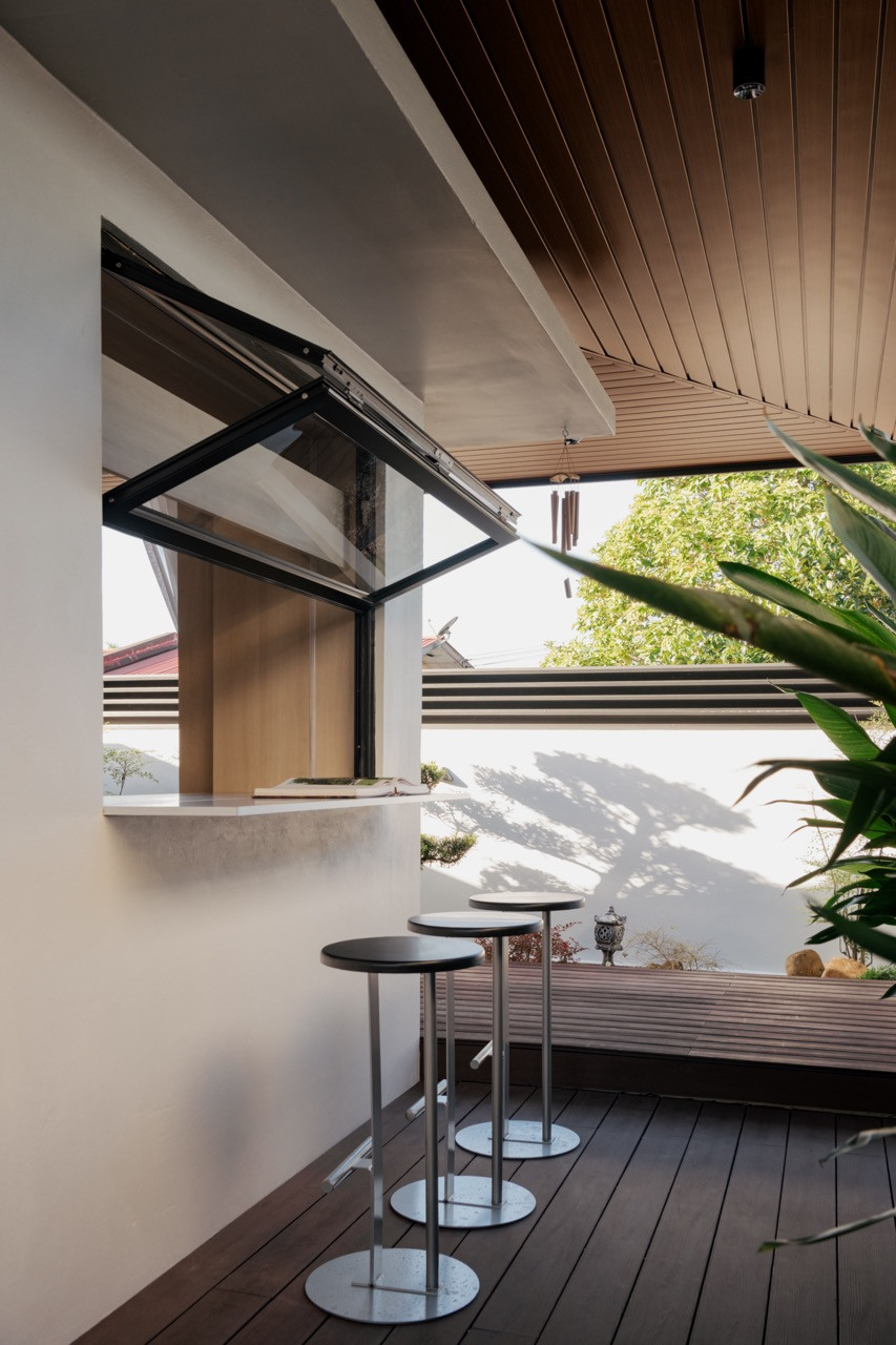 The outdoor bar counter with a fold-up pass-through window connecting to the wet kitchen, with high stools on the timber deck and garden greenery beyond