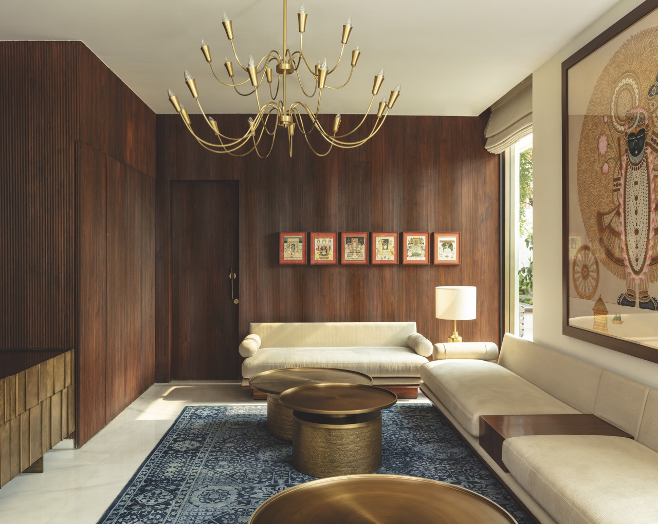 A living room with teak-panelled walls, a brass chandelier and pichwai artworks