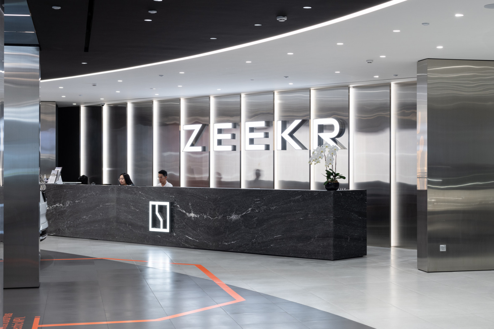 A black marble reception counter set against brushed stainless steel panels and orange floor markings establishes the high-tech brand identity of the Zeekr Centre Singapore, designed by Aarticle, ARTG Design Group's commercial design arm