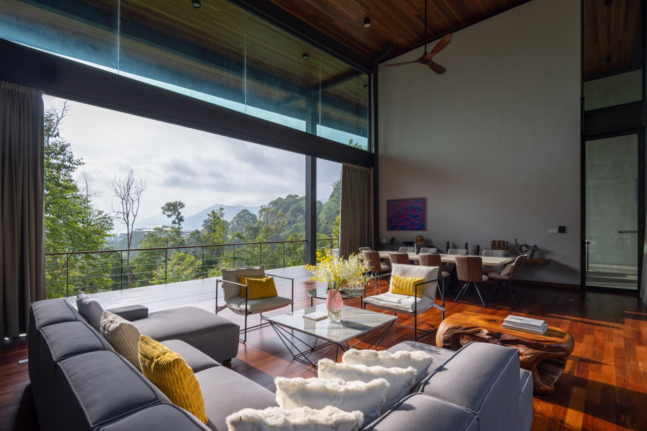The upper-level living room, with floor-to-ceiling glazing framing an unobstructed view of the forested Janda Baik hills, with an open-plan layout flowing through to the dining area behind