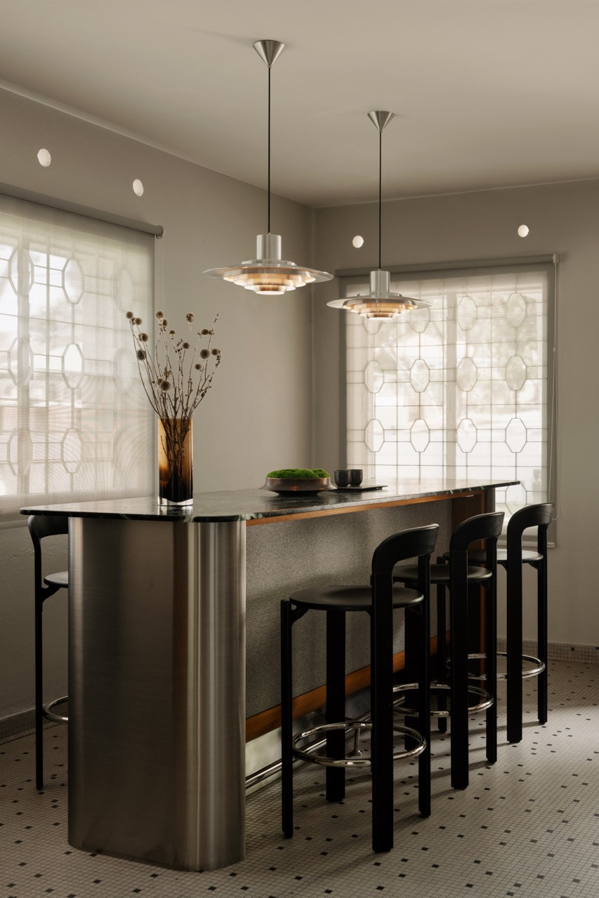The bar counter, with a dark stone top and brushed stainless steel base, sits beneath two PH5-style pendant lights in chrome