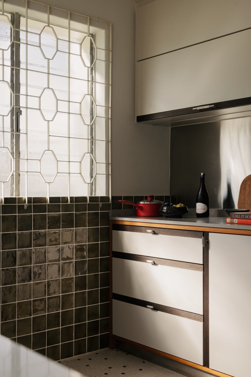 The kitchen retains its original octagonal metal window grille alongside custom cabinetry in cream and walnut, with a grey stone countertop and dark square wall tiles