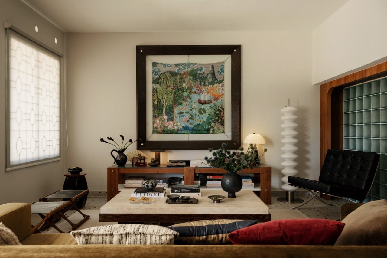 The living room looking toward the main artwork wall: the large painting in its dark timber frame, the console below stacked with objects and books, the travertine coffee table, the Barcelona chair and a stacked floor lamp