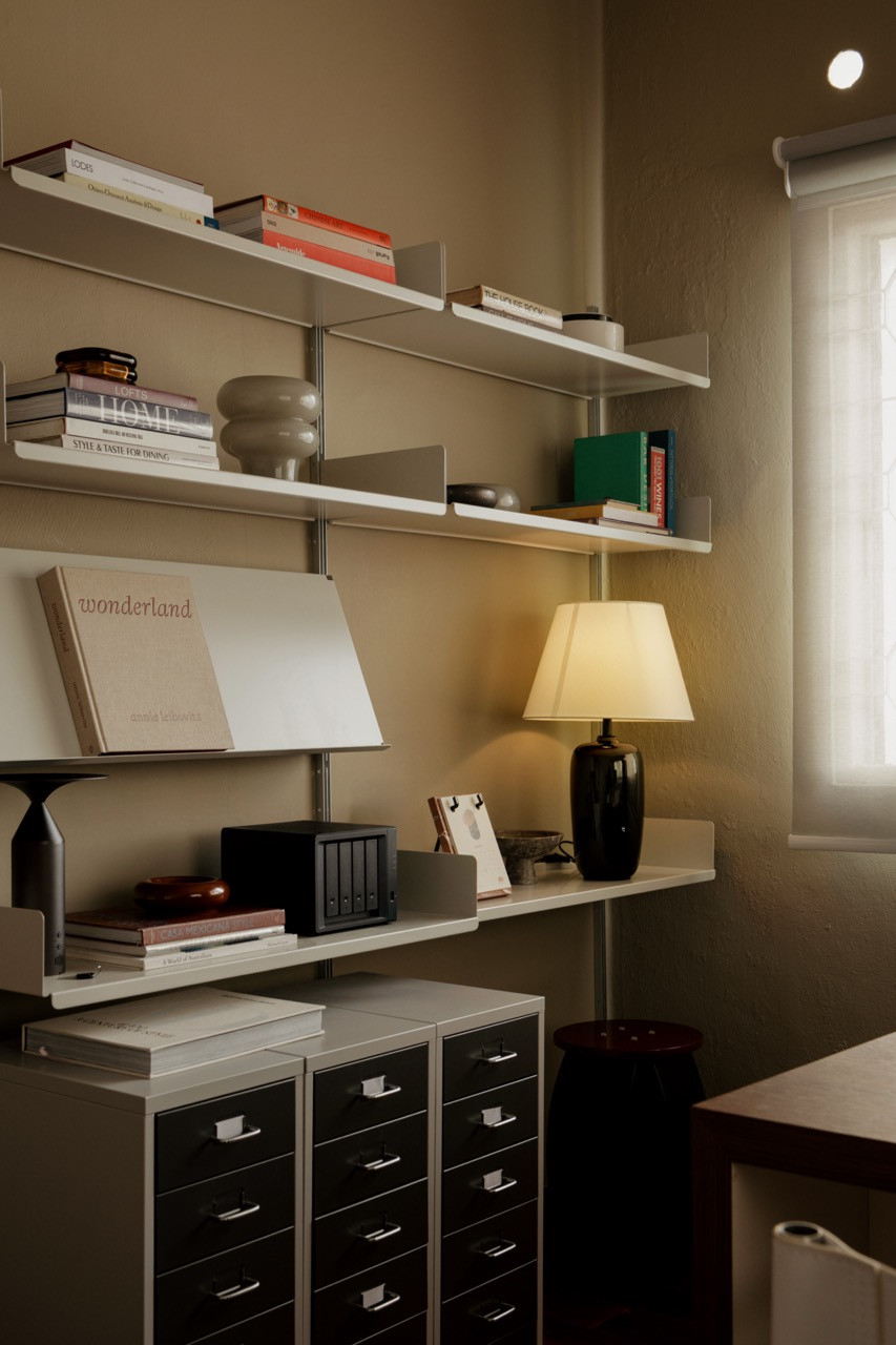 A wall-mounted shelving system in the studio holds a mix of design books, objects, and a ceramic table lamp