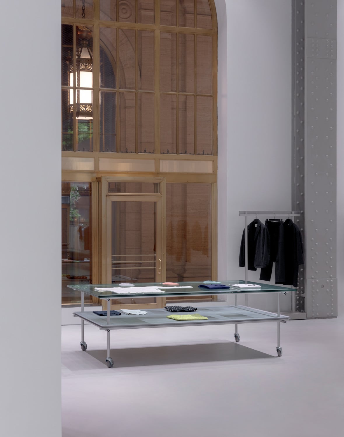 Folded garments are displayed on a two-tier glass-and-steel trolley positioned near a riveted structural column (Photo: Naho Kubota)