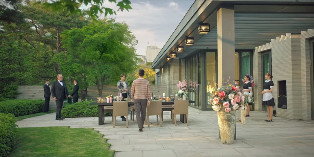 A still from The Penthouse: War in Life, filmed on the garden terrace where the stone-paved outdoor dining area and floor-to-ceiling glazing of the main living volume are visible behind the cast (Photo: SBS South Korea)