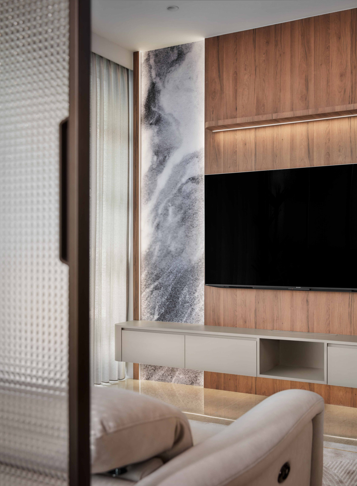 A veined marble feature wall sits beside the walnut‑panelled television console, catching the light from the window