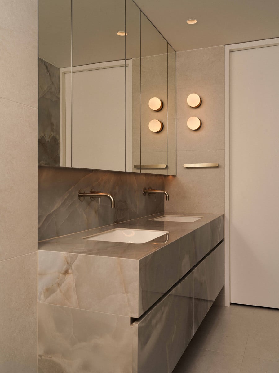 A double vanity in onyx-look stone is lit by round wall sconces reflected in the mirrored cabinetry