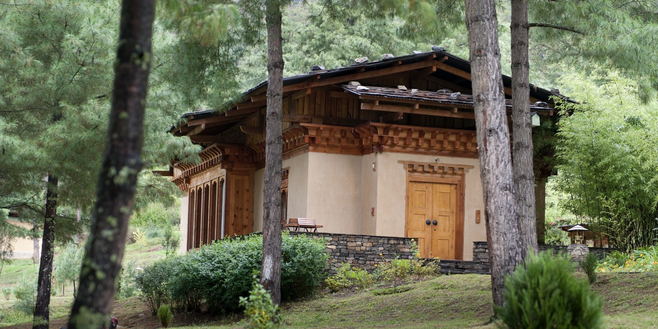 A guest villa at Uma Paro sits framed by pine and bamboo, its stone base and carved timber doorway echoing traditional Bhutanese construction