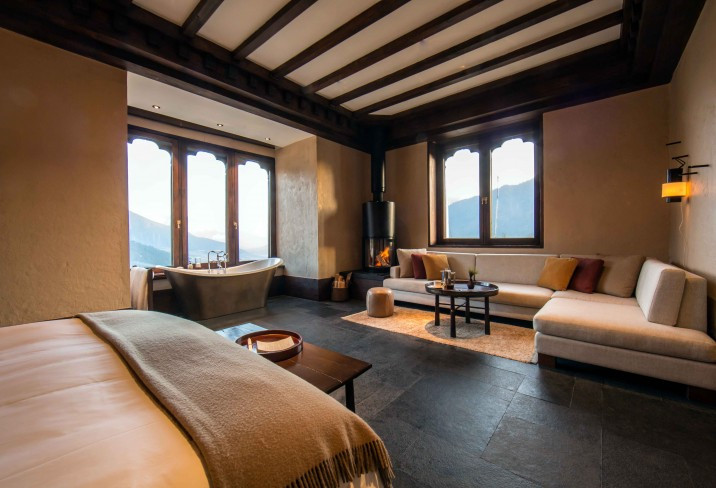 A suite at Gangtey Lodge pairs a freestanding tub and wood-burning stove beneath a dark timber-beamed ceiling, with arched windows framing the valley