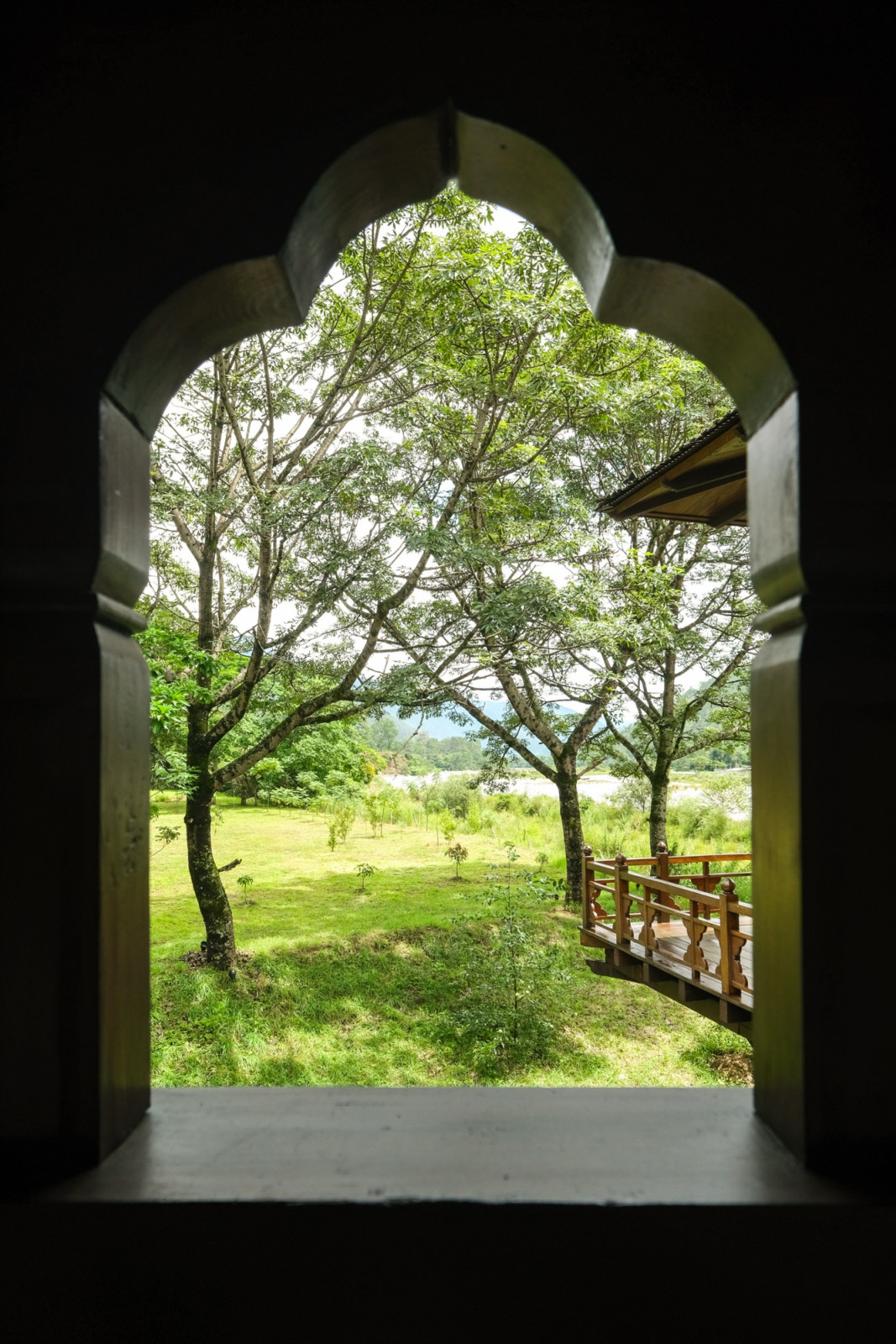 An arched window at Pemako Punakha frames a footbridge and orchard leading toward the Mo Chu River