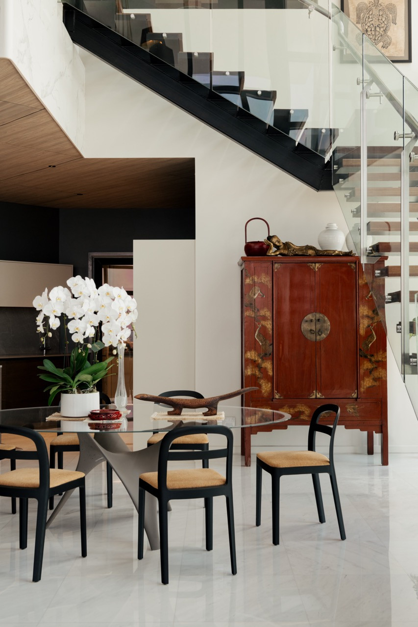 The dining area’s glass-top table and black-framed chairs sit beneath the floating staircase