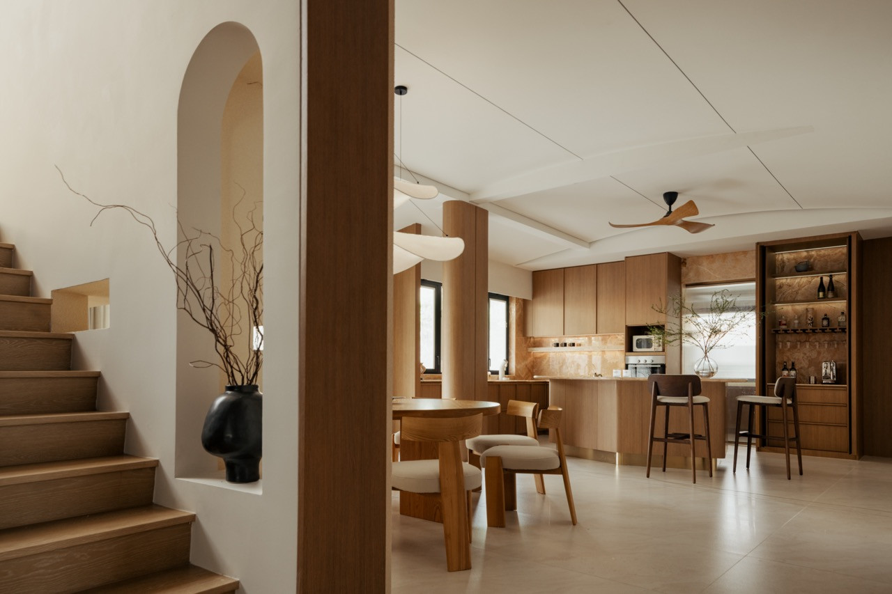 Seen from the staircase, the kitchen, dry bar, and dining area unfold as a single continuous volume beneath the home’s curved ceiling