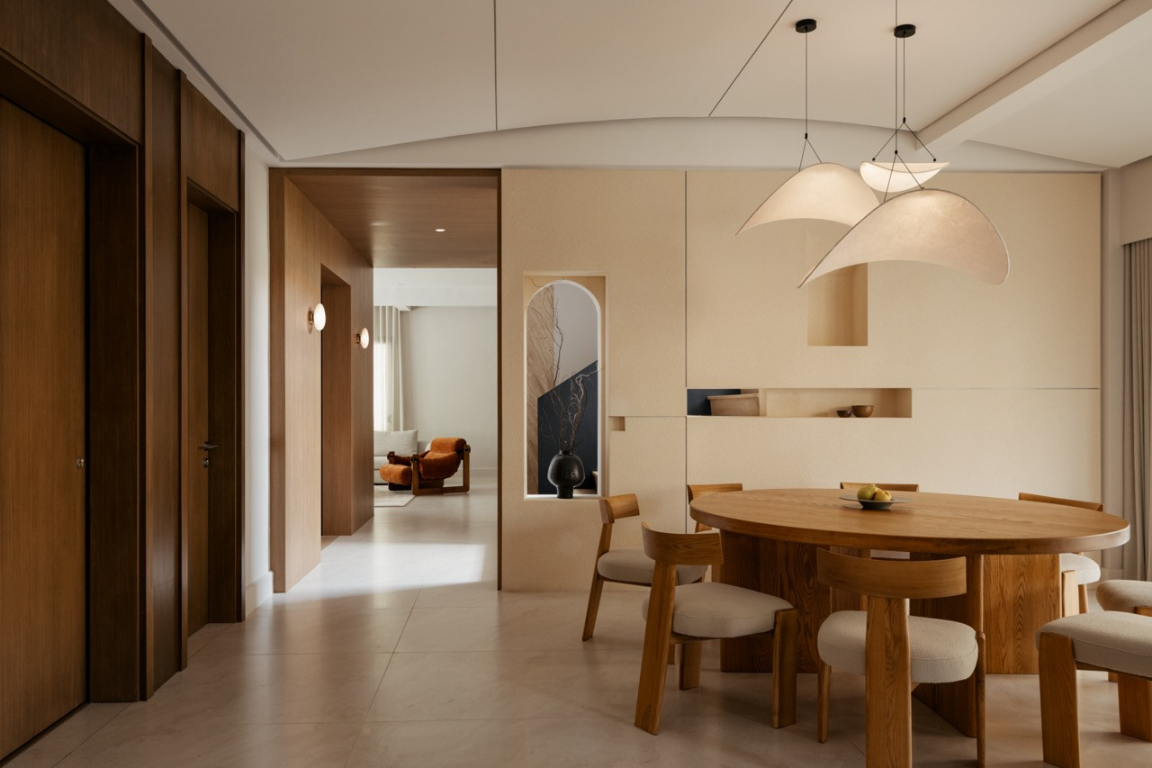 The dining area opens toward the living room beyond, framed by an arched wall niche and a sculptural double pendant