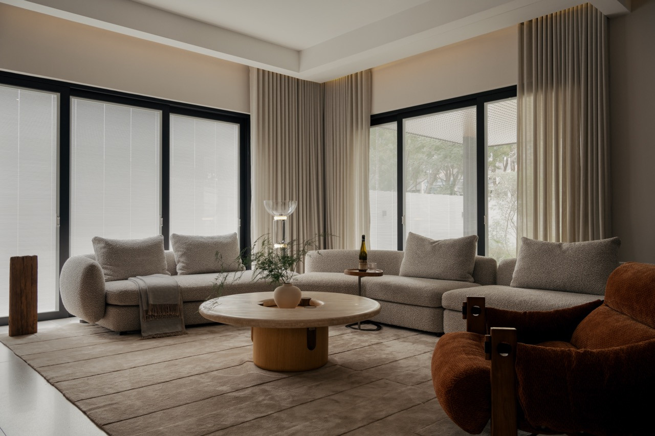 The living room is furnished with a bouclé sectional, a travertine coffee table, and a burnt-orange lounge chair, set against full-height curtains