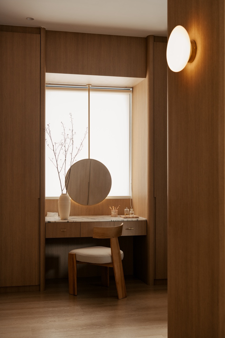 The dressing table sits within a timber alcove, a round mirror suspended above a marble-topped counter and a single ceramic vase