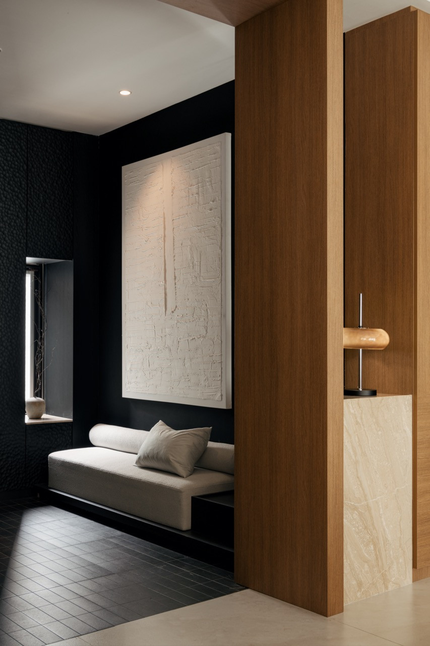 The darkened foyer pairs a textured black wall with a built-in daybed and a large tactile artwork, a slot window admitting a narrow line of daylight