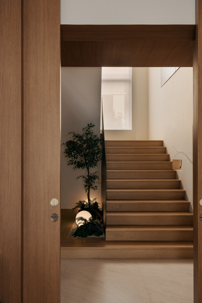 A timber staircase rises beside a small indoor planting nook, lit by a spherical floor lamp set among ferns and stones