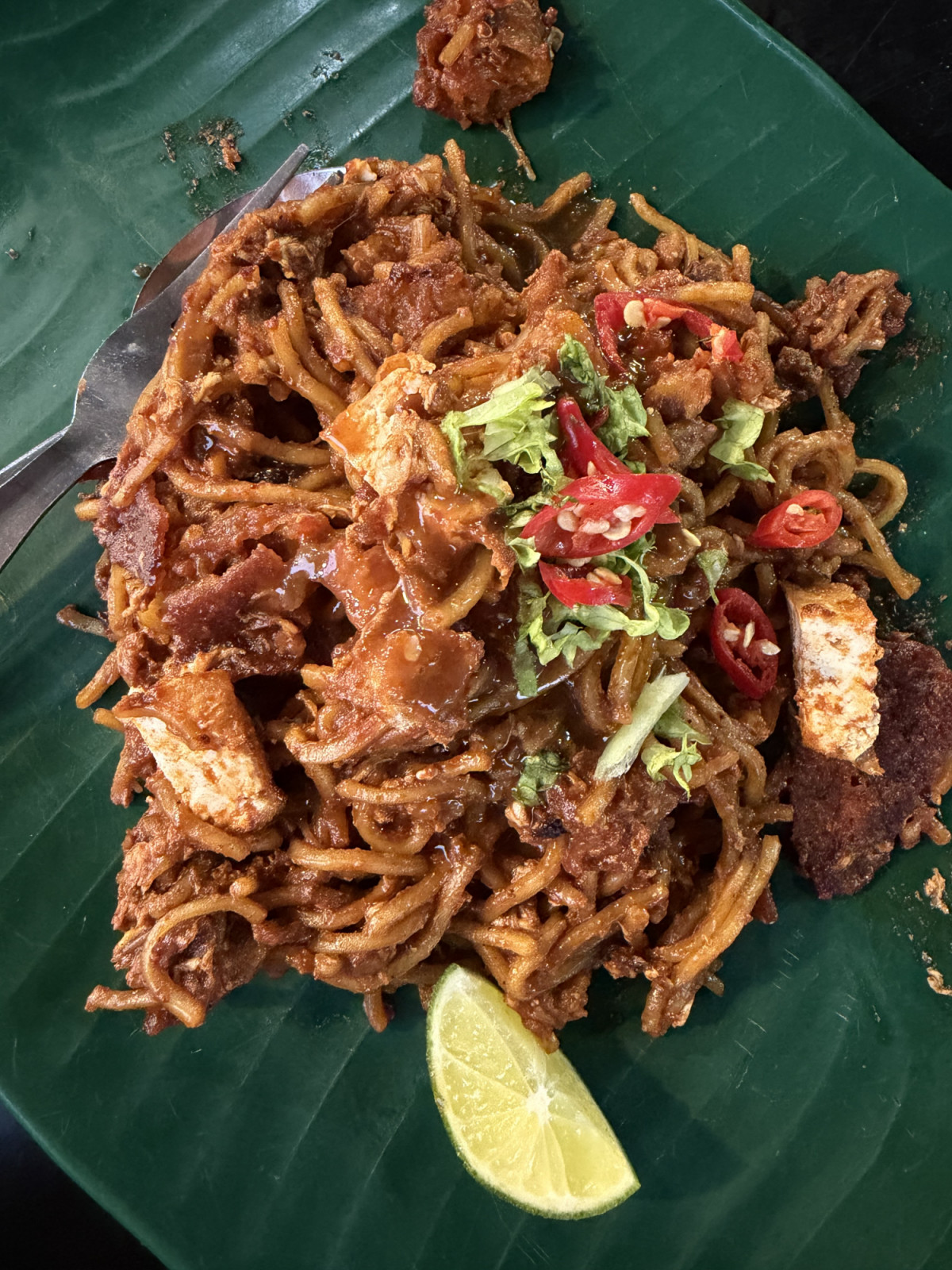 A vertical, high-angle overhead photograph from file 46519B31-4007-434C-8BCB-B7CF539CB4B8.jpg capturing a classic plate of Malaysian street food. The composition centers on a portion of Mee Goreng Mamak (Indian-Muslim style fried yellow noodles) coated in a thick, savory, and spicy reddish-brown sauce. The noodles are tossed with fried tofu cubes, bean curd fritters, eggs, and shredded cabbage, then garnished with chopped green lettuce and freshly sliced red chilies. A single fresh lime wedge rests at the b