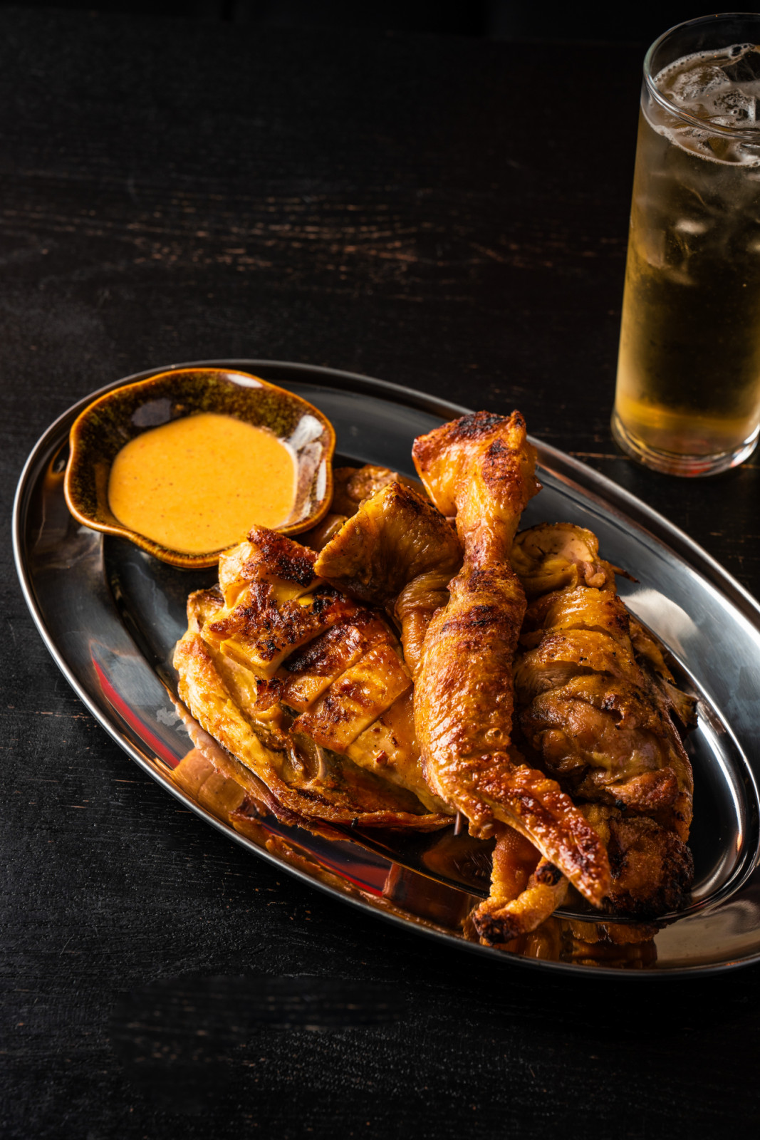 Grilled chicken pieces are presented on a metal platter with a rich-hued dipping sauce and an iced highball.