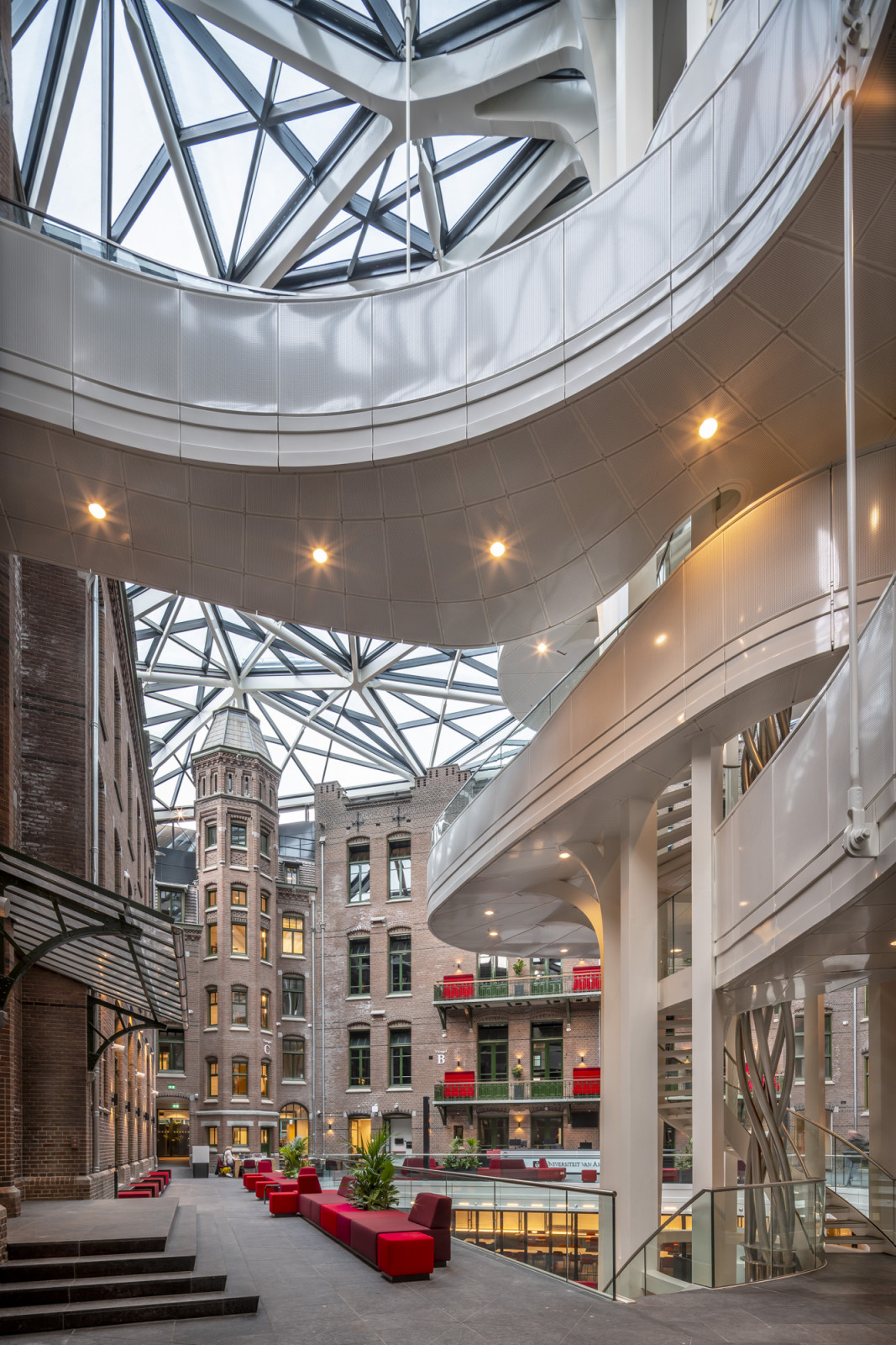 A curved white walkway sweeps past a brick corner tower original to the historic complex, its windows lit against the atrium’s steel roof structure (Photo: Jannes Linders)