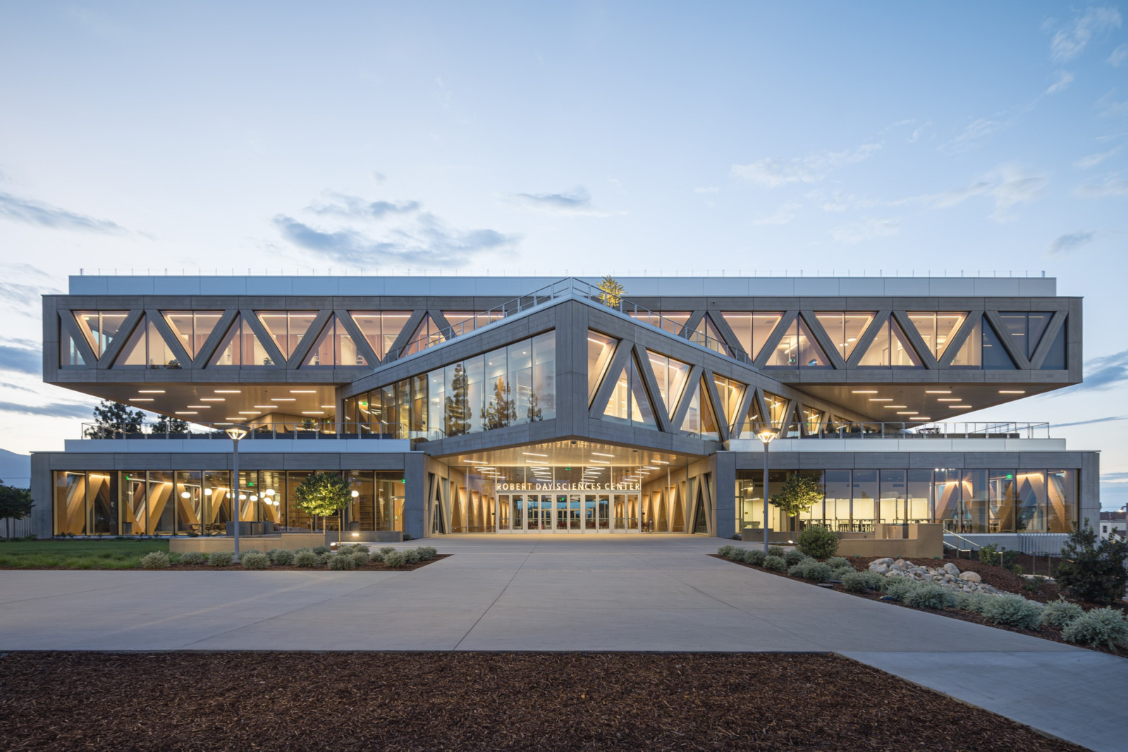 At dusk, warm light fills the timber-lined interior visible through the entrance’s angled glass volume, flanked by triangulated concrete bracing on either side (Photo: Laurian Ghinitoiu)