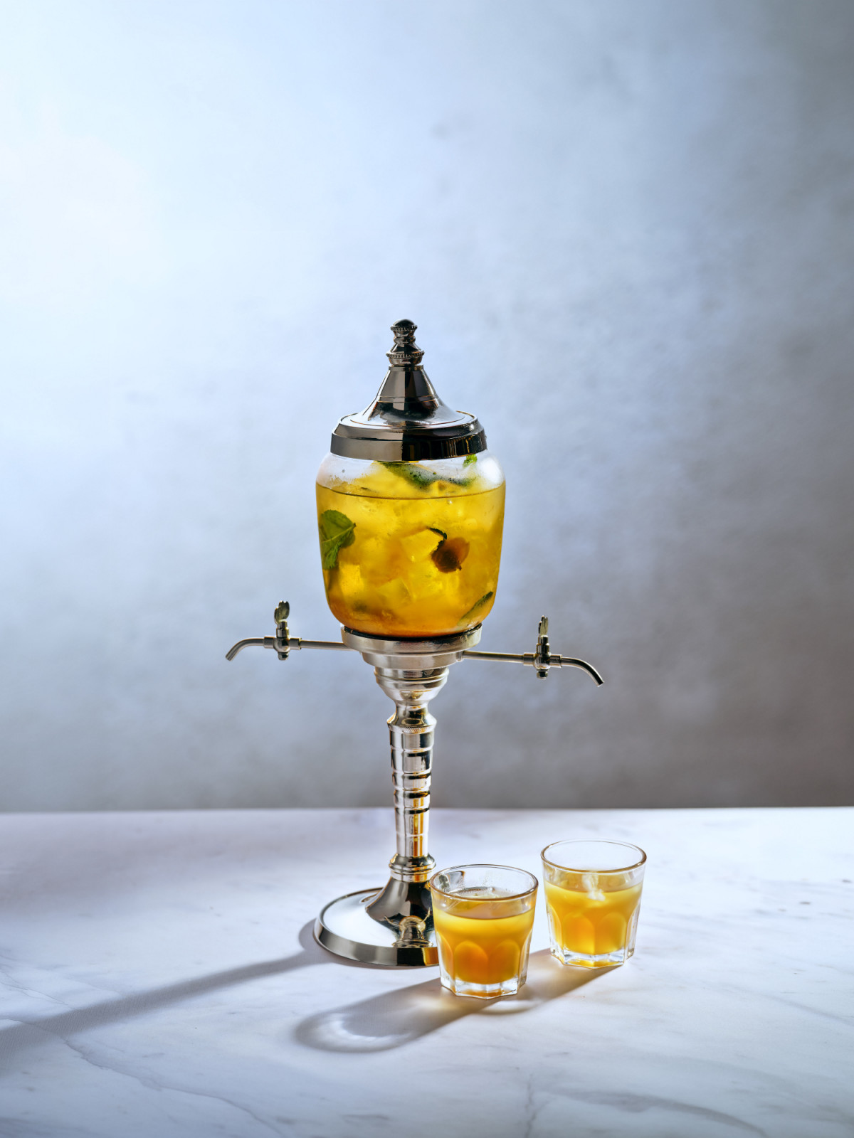 An ornate beverage dispenser pours a fruit and mint-infused cocktail into shot glasses.