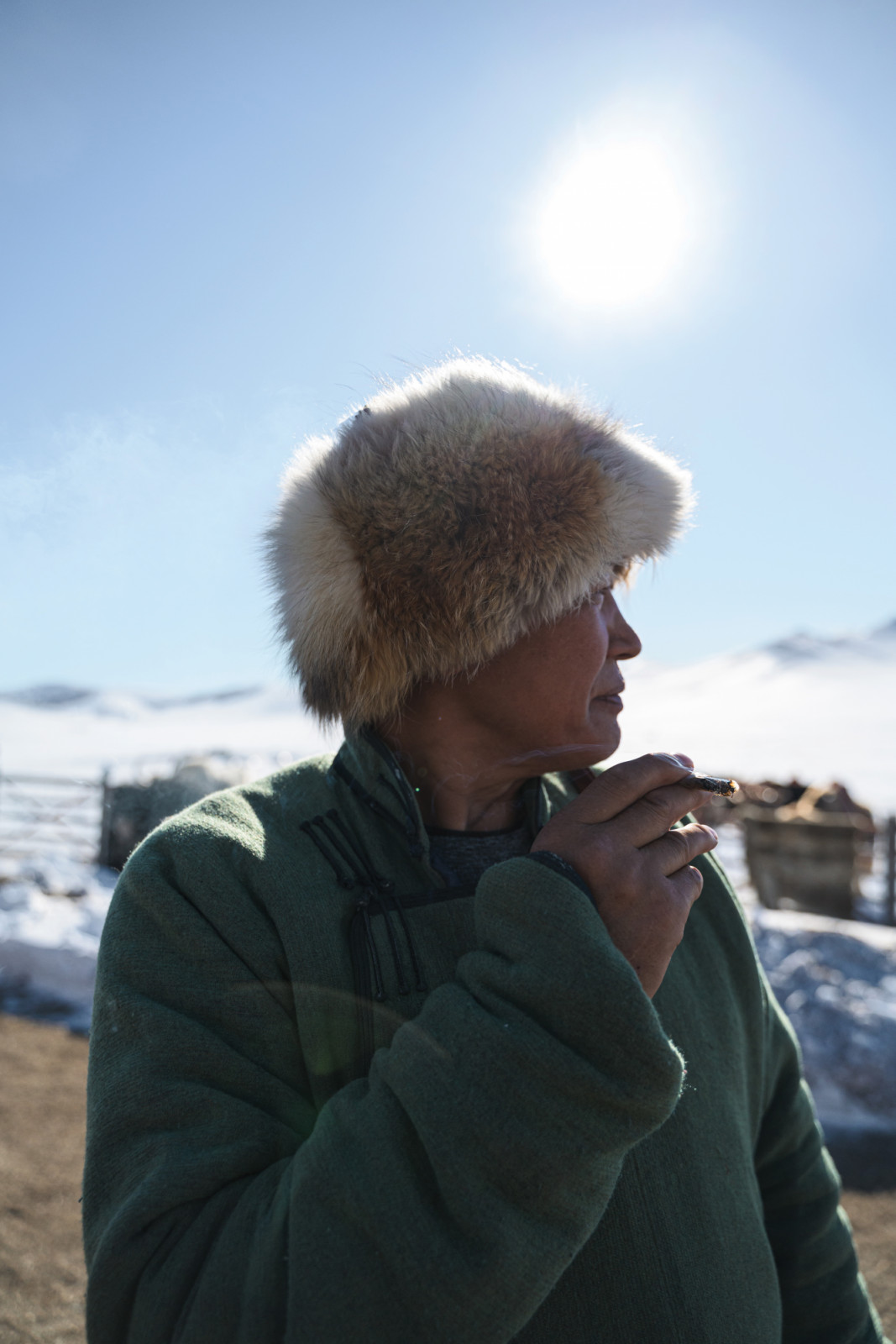 A person in a fur hat and traditional green clothing pauses with a pipe in a snowy, mountainous landscape. (Photo by Gab Mejia)