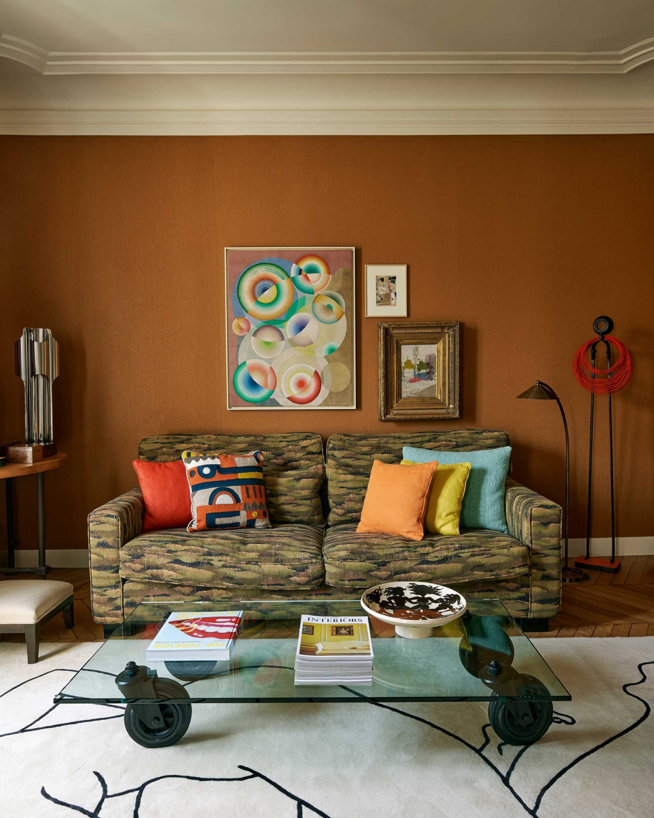 The TV room featuring a Pierre Frey sofa, a painting by Léon Texier, a Gae Aulenti glass coffee table on wheels and a Pierre Frey rug designed by Louise Bourgoin