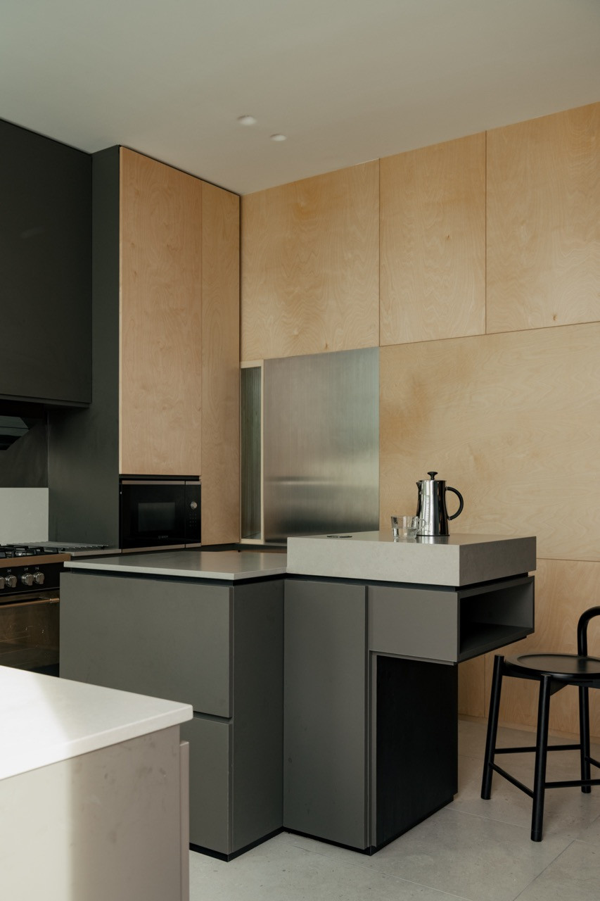 A kitchen island in matte grey cabinetry, set against birch plywood wall panelling and a stainless steel splashback