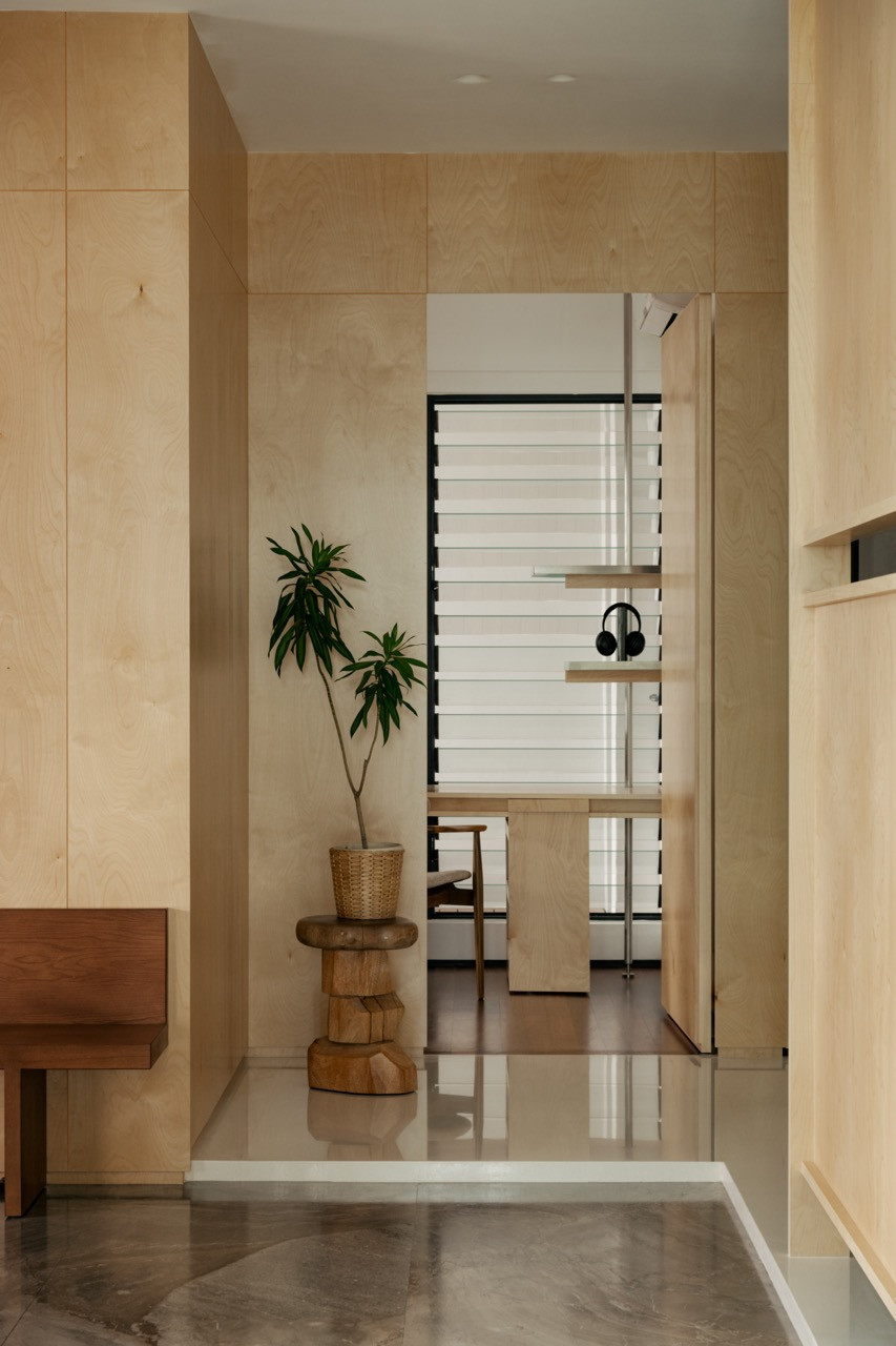 A plywood-lined entryway with a potted plant on a carved timber stool, opening onto a louvred window