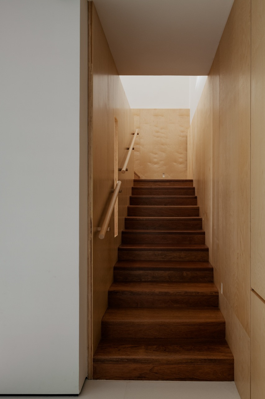 A narrow staircase finished in dark timber treads, framed by plywood-lined walls and lit from above