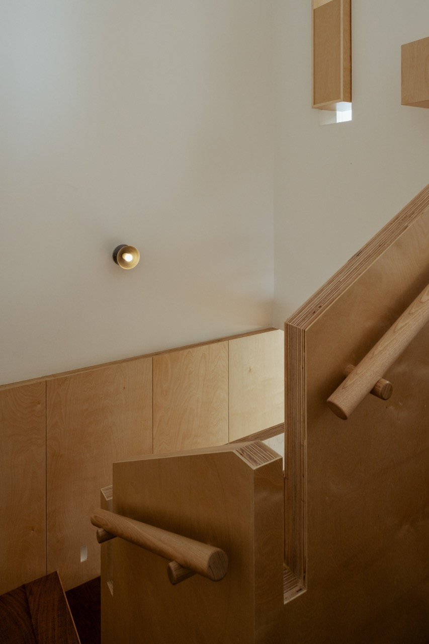 A stairwell in pale plywood, with a rounded timber handrail and a small wall-mounted light fixture