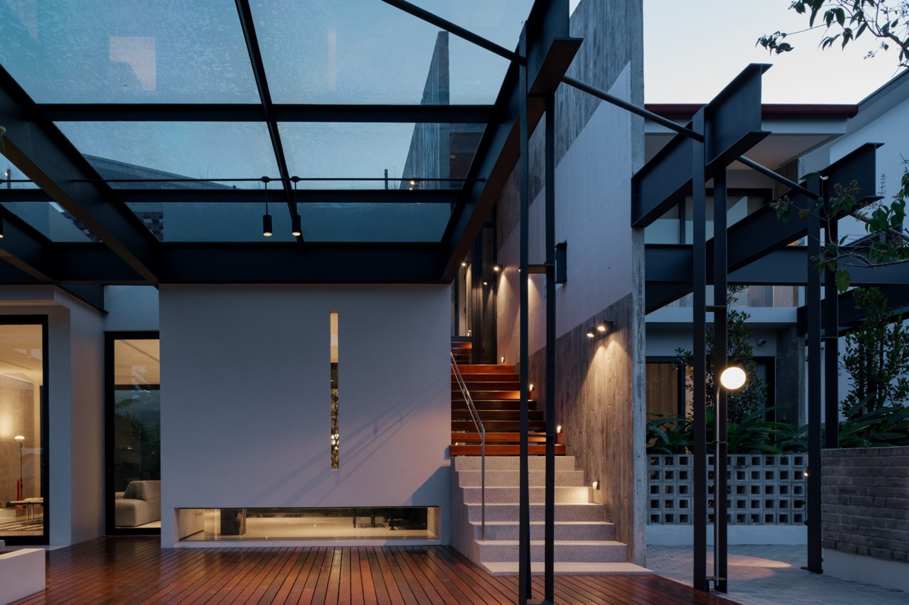 A rear deck at Habitus, Melaka, with a glass roof braced on black steel beams above a staircase in concrete and timber