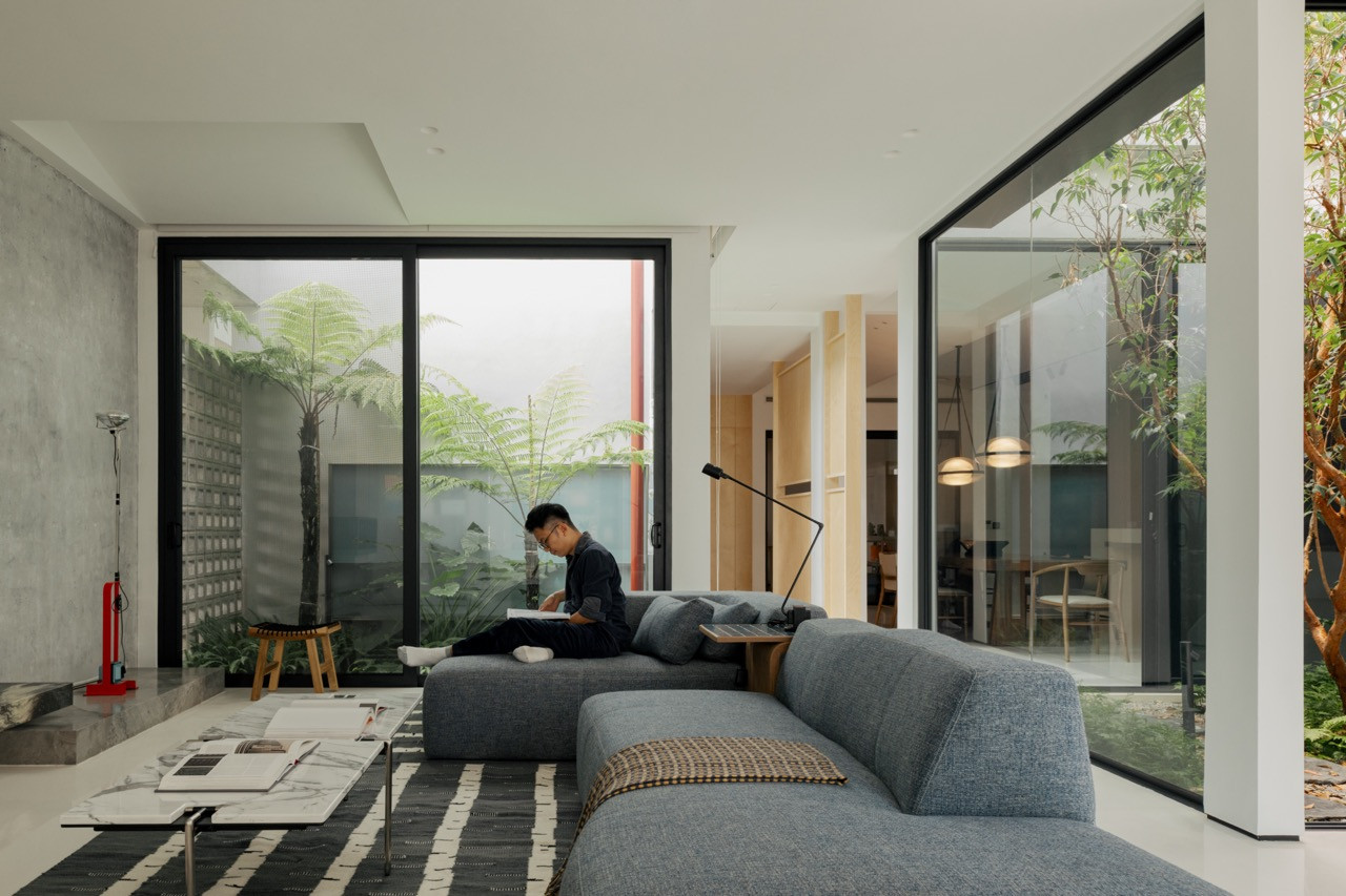 A figure reading on the living room sofa, framed by glass walls opening onto planted courtyards on two sides